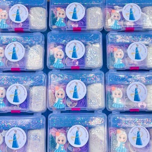 May include: Multiple clear plastic boxes filled with craft supplies. Each box contains a small doll, glitter, and decorative elements. The boxes are decorated with stickers featuring a princess and the text "Thank you for celebrating with me."