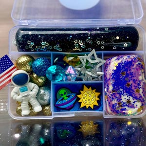 May include: A clear plastic container filled with various space-themed items, including blue and gold glitter slime, blue and gold beads, a small astronaut figurine, a green alien figurine, a yellow sun, and a blue and white galaxy slime.