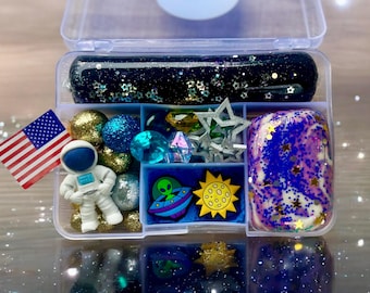 May include: A clear plastic container filled with various space-themed items, including blue and gold glitter slime, blue and gold beads, a small astronaut figurine, a green alien figurine, a yellow sun, and a blue and white galaxy slime.
