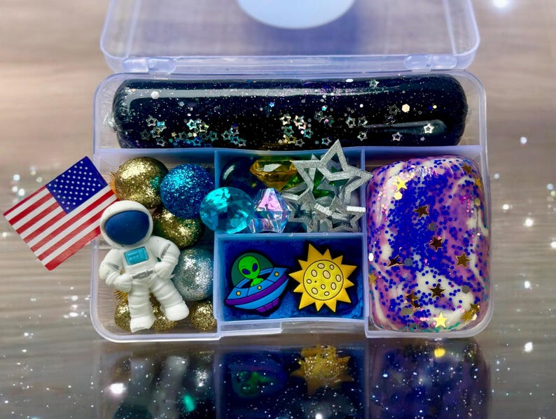 May include: A clear plastic container filled with various space-themed items, including blue and gold glitter slime, blue and gold beads, a small astronaut figurine, a green alien figurine, a yellow sun, and a blue and white galaxy slime.