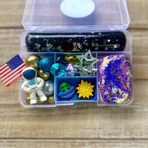 May include: A clear plastic container with compartments filled with space-themed slime, charms, and toys. The slime is black with glitter and stars, and the charms include an astronaut, a UFO, a moon, and a star. There are also blue and gold beads, and a small American flag.