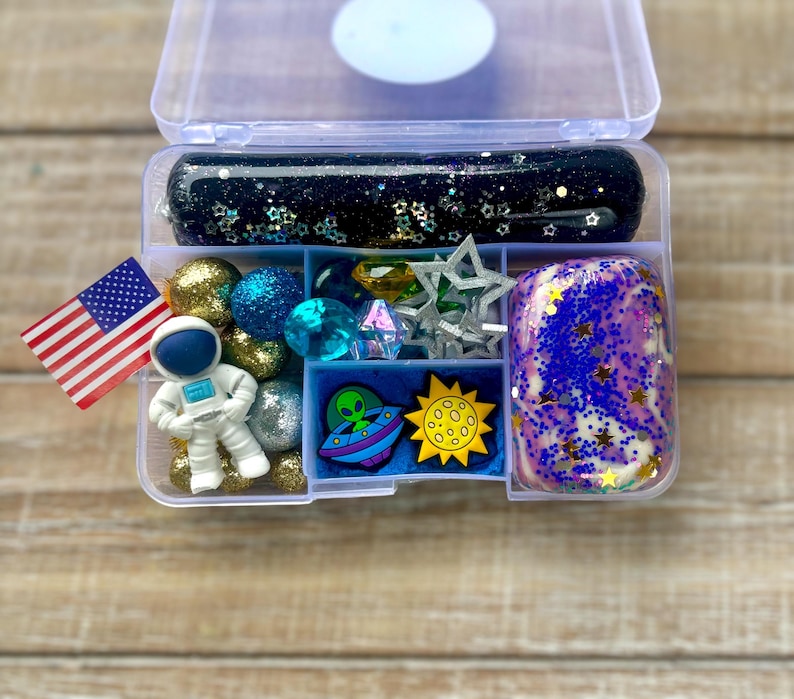 May include: A clear plastic container with compartments filled with space-themed slime, charms, and toys. The slime is black with glitter and stars, and the charms include an astronaut, a UFO, a moon, and a star. There are also blue and gold beads, and a small American flag.