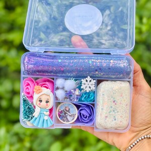 May include: A clear plastic box with a pink clasp, filled with various craft supplies. Includes glittery slime, miniature figurines, pink and purple roses, a snowflake, and other decorative elements. The box is held against a blurred green background.