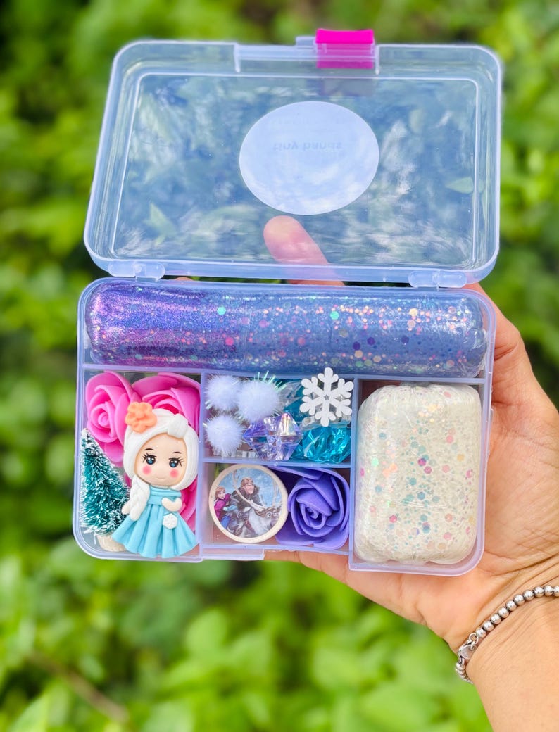 May include: A clear plastic box with a pink clasp, filled with various craft supplies. Includes glittery slime, miniature figurines, pink and purple roses, a snowflake, and other decorative elements. The box is held against a blurred green background.