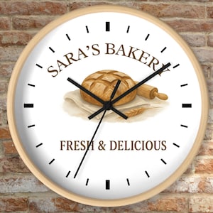 May include: A round wall clock with a light wood frame. The clock face features the text "Sara's Bakery" above a watercolor illustration of a loaf of bread, rolling pin, and wheat. The words "Fresh & Delicious" are below the illustration.