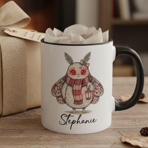 May include: White ceramic mug with a black handle and rim, featuring a cartoon moth illustration with a red and white scarf, holding a lantern. The name "Stephanie" is written below the moth. The mug is filled with white tissue paper.