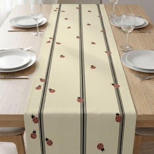 May include: A cream-colored table runner with a repeating pattern of red ladybugs and black vertical stripes. The runner is placed on a wooden table set with white plates, silverware, and wine glasses.