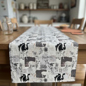 May include: A farmhouse-style table runner with a cream background and a repeating pattern of black and white cows, chickens, goats, and sunflowers. The words "Farm House" are printed on the runner. The runner is placed on a wooden table.