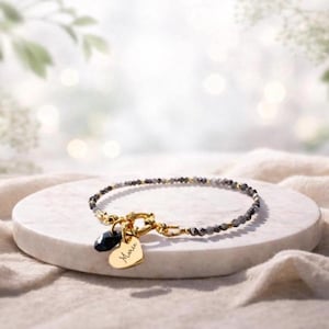 Könnte beinhalten: Armband mit grauen und schwarzen Perlen, goldenem Verschluss und Anhängern. Das Armband hat kleine Perlen mit goldenen Details. Ein schwarzer tropfenförmiger Anhänger und ein goldener Herzanhänger mit dem Namen "Marie" sind befestigt.