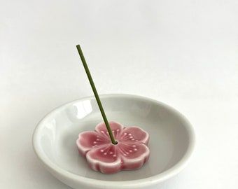 May include: A white ceramic incense holder shaped like a pink flower with a green incense stick standing upright in the centre.