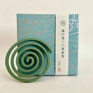 Puede incluir: Una espiral de mosquito verde con una caja de patrón azul y plateado. La caja tiene texto japonés que dice "Incienso de aroma marino".