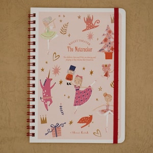 Puede incluir: Una libreta de espiral rosa y blanca con cierre elástico rojo. La portada presenta ilustraciones de personajes del ballet El Cascanueces, incluyendo a Clara, Fritz, el Cascanueces y un unicornio. El texto "Ballet Theater The Nutcracker" está impreso en la portada.