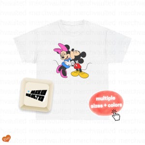May include: White t-shirt featuring a colorful graphic of Mickey Mouse and Minnie Mouse embracing. The design includes the text "multiple sizes + colors". The shirt is a classic crew neck.