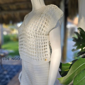 Puede incluir: Conjunto de top y pantalones cortos de crochet blanco con cuello en V y mangas cortas. El top tiene un tejido suelto y abierto y los pantalones cortos son un poco más cortos. El conjunto está hecho de una tela suave y ligera, perfecto para el clima cálido.