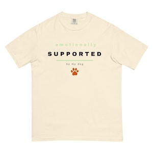May include: Cream-colored t-shirt with the text "emotionally SUPPORTED by my dog" in black and green. A brown paw print graphic is below the text. The shirt has a crew neck and short sleeves.