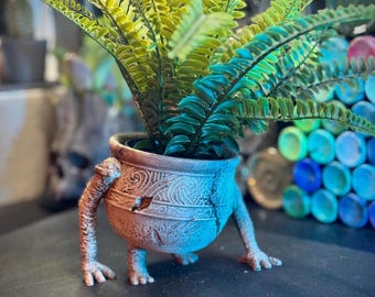 May include: A decorative planter with a fern. The planter is a light brown color with a detailed pattern and legs. The fern has green and yellow leaves. The planter is on a dark surface.