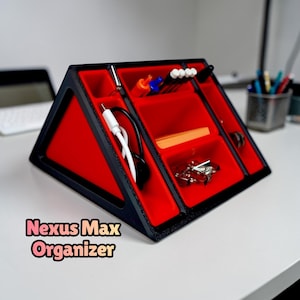 Puede incluir: Un organizador de escritorio negro y rojo con múltiples compartimentos. El organizador triangular contiene bolígrafos, un cable de carga y otros pequeños útiles de oficina. El texto "Nexus Max Organizer" se muestra en la parte inferior.