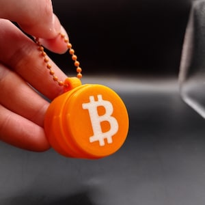 Könnte beinhalten: Orangener Schlüsselanhänger mit einem weißen Bitcoin-Symbol auf einem runden Anhänger.