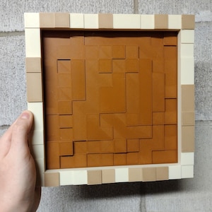 Könnte beinhalten: Ein quadratisches 3D-Wandkunstwerk mit einem braunen, strukturierten Mittelteil und einem cremefarbenen Rahmen. Der Rahmen hat abwechselnd beige und cremefarbene quadratische Blöcke. Das Kunstwerk wird von einer Hand vor einer grauen Backsteinmauer gehalten.