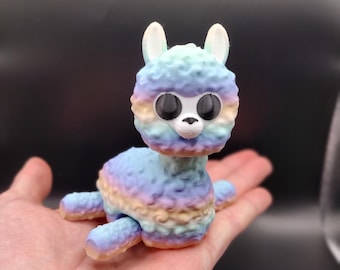 Könnte beinhalten: Ein kleines, farbenfrohes, 3D-gedrucktes Lama-Spielzeug mit großen Augen. Das Lama liegt mit ausgestreckten Beinen. Das Lama besteht aus einem weichen, strukturierten Material und ist in Blautönen, Rosa und Gelb bemalt.