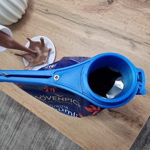 May include: A blue plastic bag dispenser with a handle and a spout. The dispenser is attached to a bag of "M&Ouml;VENPICK Crema Cumi" coffee.