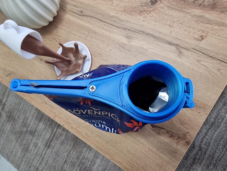 May include: A blue plastic bag dispenser with a handle and a spout. The dispenser is attached to a bag of "M&Ouml;VENPICK Crema Cumi" coffee.
