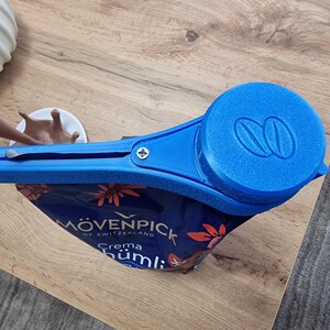 May include: Blue plastic bag clip with a screw and a round blue cap with a double coffee bean logo. The clip is attached to a blue and red bag with the text "M&Ouml;VENPICK OF SWITZERLAND Crema H&uuml;mlı" printed on it.