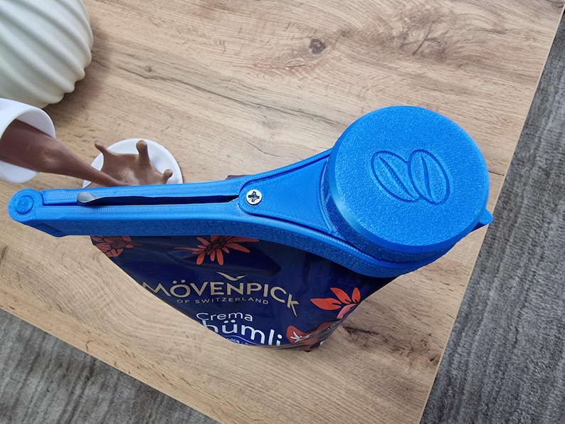 May include: Blue plastic bag clip with a screw and a round blue cap with a double coffee bean logo. The clip is attached to a blue and red bag with the text "M&Ouml;VENPICK OF SWITZERLAND Crema H&uuml;mlı" printed on it.