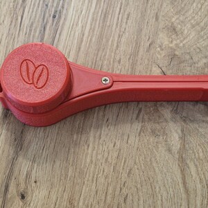 May include: A red plastic coffee scoop with a hinged handle and a round, removable container with a coffee bean logo.