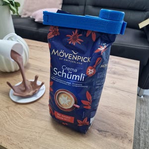 May include: A blue plastic bag clip is holding a blue and red bag of M&ouml;venpick Crema Sch&uuml;mli coffee. The bag has a picture of a cup of coffee with a star-shaped design on top. The text on the bag reads "M&ouml;venpick of Switzerland Crema Sch&uuml;mli 100% Hochland Arabica Sorgzame Langzeitrostung 1000g 1000".