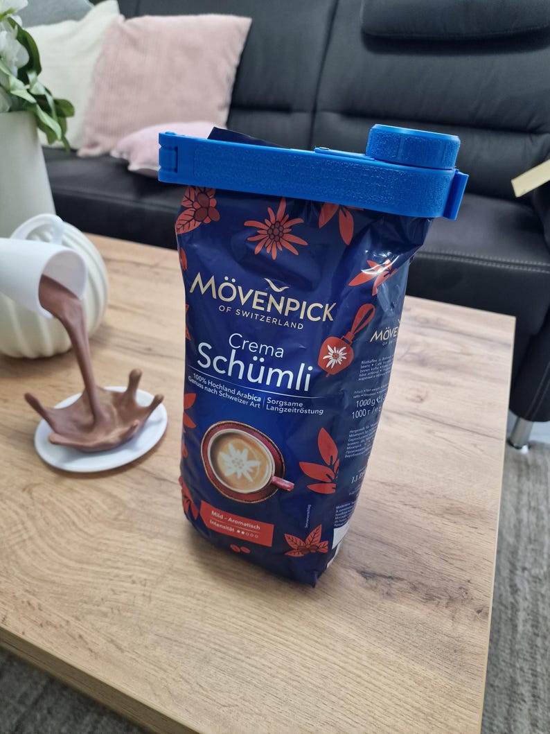 May include: A blue plastic bag clip is holding a blue and red bag of M&ouml;venpick Crema Sch&uuml;mli coffee. The bag has a picture of a cup of coffee with a star-shaped design on top. The text on the bag reads "M&ouml;venpick of Switzerland Crema Sch&uuml;mli 100% Hochland Arabica Sorgzame Langzeitrostung 1000g 1000".