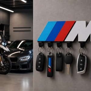 Peut inclure: Un porte-clés mural avec le logo BMW en bleu, rouge et blanc. Il comporte six crochets avec différentes clés de voiture. L'arrière-plan montre une moto et une voiture gris foncé.