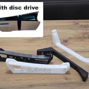 Può includere: Una collezione di supporti per console stampati in 3D in nero, bianco e grigio. I supporti presentano un design geometrico unico. L'immagine include anche una scatola bianca con la scritta "with disc drive" e una console.