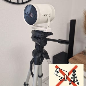 May include: A white cylindrical projector mounted on a black tripod with a custom mount. The tripod has silver legs. A sign indicates that the projector and tripod are not included. The background includes a wall clock and a black speaker.