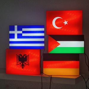 Könnte beinhalten: Beleuchtete rechteckige Tafeln mit den Flaggen von Griechenland, Albanien, der Türkei, Palästina und Deutschland. Die Flaggen sind von innen beleuchtet und erzeugen eine lebendige Darstellung nationaler Symbole. Die Tafeln sind auf einem weißen Regal angeordnet.
