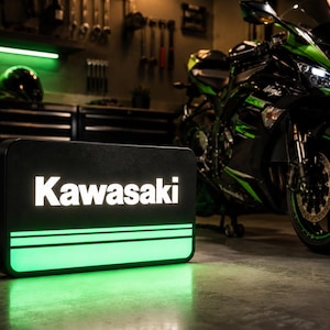 May include: Black rectangular sign with the word "Kawasaki" in white, illuminated with green light. A green and black motorcycle is parked in the background. A black helmet is on a shelf in the background.