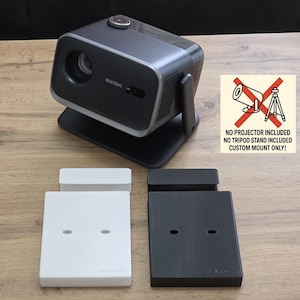May include: A black and silver projector with a custom mount, and two rectangular mounts, one white and one black. The image includes a sign that says "NO PROJECTOR INCLUDED, NO TRIPOD STAND INCLUDED, CUSTOM MOUNT ONLY!"