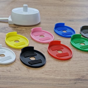 Könnte beinhalten: Eine weiße Ladestation für eine elektrische Zahnbürste mit acht verschiedenen farbigen Zahnbürstenhaltern. Die Halter sind in den Farben rosa, gelb, weiß, schwarz, blau, rot, grün und grau.