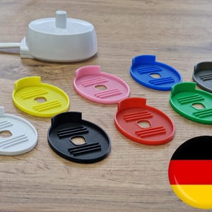 Könnte beinhalten: Eine weiße elektrische Ladestation mit Netzkabel und acht ovalen Kunststoffabdeckungen in verschiedenen Farben wie Gelb, Pink, Blau, Grau, Grün, Rot, Schwarz und Weiß, jeweils mit einer mittigen Aussparung und erhabenen horizontalen Rillen.