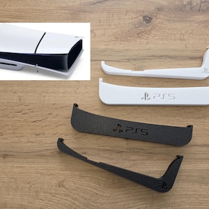 Könnte beinhalten: Weiße und schwarze PlayStation 5 Konsole mit passenden weißen und schwarzen Konsolenständern. Die Ständer tragen das PlayStation-Logo und den Schriftzug "PS5". Die Konsole und die Ständer liegen auf einer Holzoberfläche.