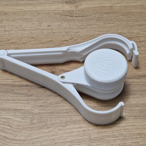 May include: White plastic tool with a hinged design. The tool has a round, white cap with a logo on it. The tool is designed to open and close containers.