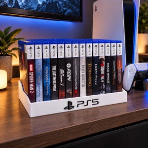 Pode incluir: Um suporte de armazenamento de plástico branco para videojogos com o logótipo da PS5 na parte frontal. O suporte contém uma fila de treze caixas de jogos da PlayStation 5, incluindo títulos como Spider-Man 2, God of War e Elden Ring.