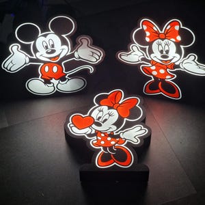 May include: Three black and white cartoon character figures of Mickey Mouse and Minnie Mouse. The figures are made of foam and have a white outline. The figures are illuminated from behind, creating a glowing effect. Mickey Mouse is wearing red shorts and Minnie Mouse is wearing a red dress with white polka dots.