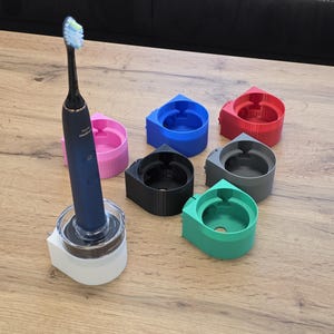 Könnte beinhalten: Eine blaue und weiße elektrische Zahnbürste mit einer Sammlung farbenfroher Zahnbürstenhalter. Die Halter sind in Pink, Blau, Rot, Schwarz, Grau und Grün gehalten. Die Zahnbürste befindet sich in einem weißen Halter.
