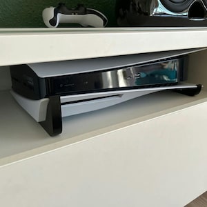 Könnte beinhalten: Eine weiße und schwarze Spielekonsole befindet sich in einer weißen Schublade. Die Konsole ist nach oben geneigt und hat eine schwarze Frontplatte mit silbernem Rahmen. Die Schublade ist geöffnet und die Konsole ist teilweise sichtbar.