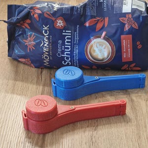 Peut inclure: Deux scelleurs de capsules de café, l'un bleu et l'autre rouge, sont posés sur une surface en bois. Les scelleurs sont en plastique et ont une poignée qui se replie. Les scelleurs servent à sceller les capsules de café après leur ouverture.