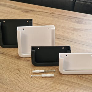 May include: Four wall-mounted storage containers in black and white, arranged on a wooden surface. Each container has a rectangular shape with a curved bottom and a hole for mounting. Two screws and anchors are also visible.