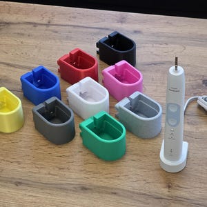 Könnte beinhalten: Eine Sammlung farbenfroher Zahnbürstenhalter in Gelb, Blau, Rot, Schwarz, Rosa, Weiß, Grau und Grün. Eine weiße elektrische Zahnbürste mit Ladestation und Kabel ist ebenfalls vorhanden. Die Halter sind zur Aufnahme der Zahnbürste konzipiert.