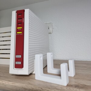 Op de afbeelding: Een witte router met een rode zijkant en een zwarte knop met het label "Connect". De router is gemonteerd op een witte wandbeugel met twee beugels. De router heeft een label aan de zijkant met de tekst "Info, Connect, Fon, WLAN, Power/Cable".