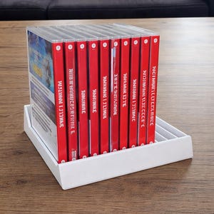 May include: A collection of video game cases with red spines, displayed in a white organizer on a wooden surface. The game cases are stacked together, with the titles facing outwards. The background includes a sofa.